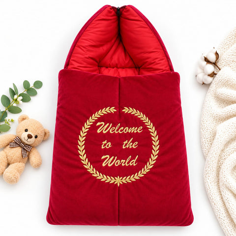 A red quilted baby sleeping bag or pouch with a hood, featuring a gold embroidered wreath and the words 'Welcome to the World' on the front. The bag is styled in a flat-lay arrangement with a teddy bear, green leaves, cotton bolls, and a cream knit blanket on a white background.