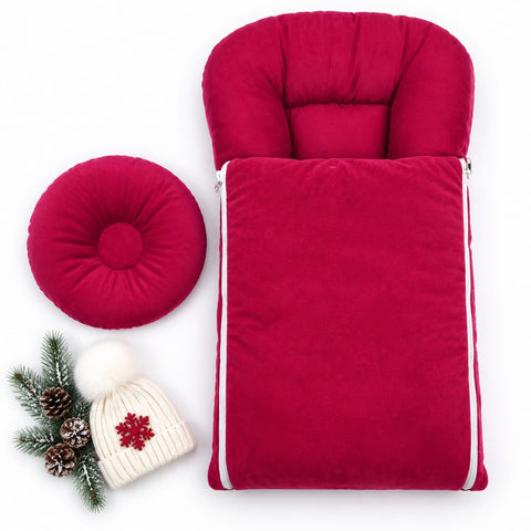 A bright pinkish-red plush baby footmuff and matching round cushion set, shown on a white background with a small holiday-themed beanie and pine sprigs as styling props.