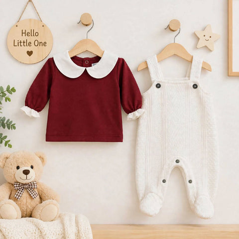 A baby clothing set displayed on wooden hangers against a light wall, featuring a long-sleeve burgundy top with a white Peter Pan collar and white ruffle trim, paired with white textured footless overalls with black buttons. A teddy bear and wooden decor hang below.