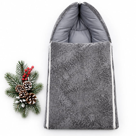 A gray plush fleece baby footmuff with a zippered closure and padded hood, displayed next to a decorative holiday pine branch with pinecones and red berries on a white background.