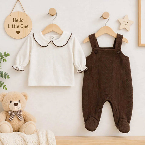 A baby outfit display on wooden hangers against a light wall, featuring a white long-sleeve top with a dark brown Peter Pan collar and cuffs, and a dark brown footed overall romper. A teddy bear sits on a blanket at the lower left, with decorative wall hangings and a small sign reading 'Hello Little One'.