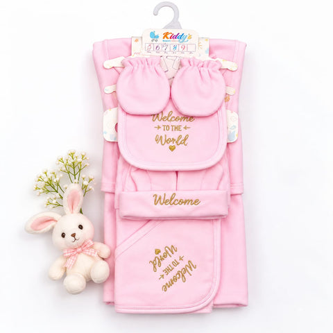 A pink baby gift set including a folded blanket, a hooded towel, a bib, and matching mittens, all printed with 'Welcome to the World' in gold lettering. The set is displayed on a white background next to a small plush bunny toy and a tiny sprig of white flowers.