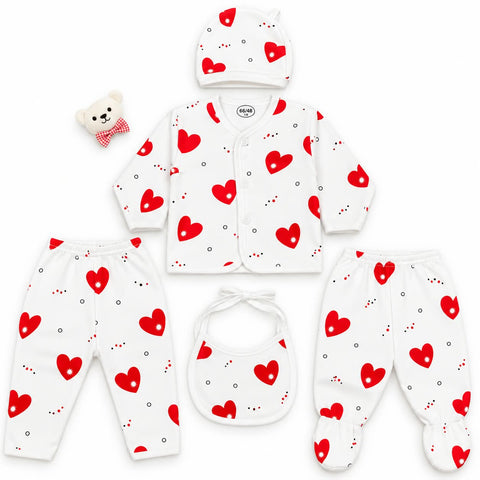 A 5-piece baby clothing set laid out on a white background, featuring a long-sleeve cardigan, footed pants, a bib, a hat, and a small teddy bear accessory. The set is white with a red heart and small black dot pattern.